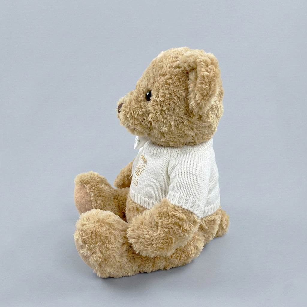 A traditional style seated teddy bear with a white knitted jumper embroidered with a crown or tiara design and the year 2026 in gold thread.