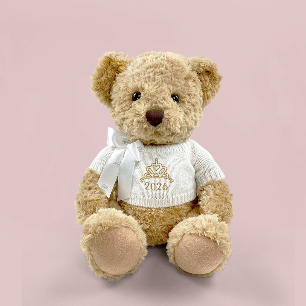 A traditional style seated teddy bear with a white knitted jumper embroidered with a crown or tiara design and the year 2026 in gold thread.